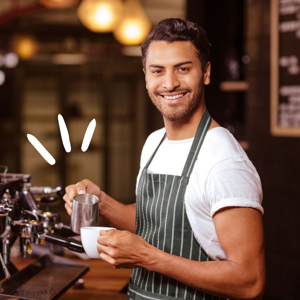 A male barista holding a milk jug and coffee cup as he makes a barista coffee.