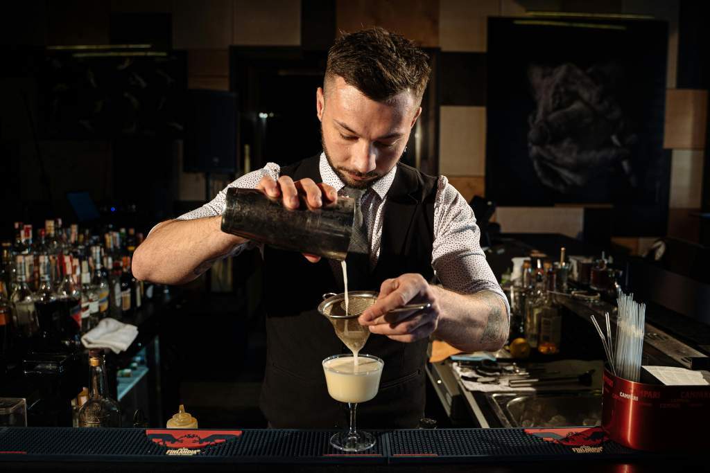A male bartender stands behind a dimly lit bar, carefully pouring a creamy cocktail through a fine strainer into a stemmed glass. He wears a white shirt with rolled sleeves, a black vest, and a focused expression. Bottles, bar tools, and mixers line the counter and shelves behind him.