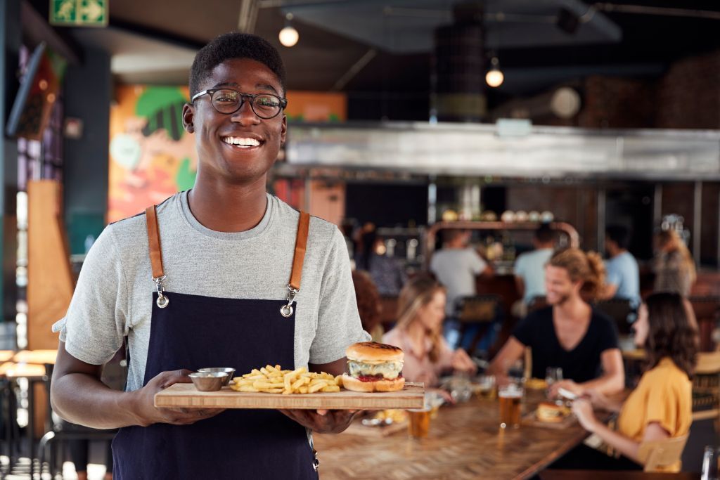 Hospitality worker in apron serving a burger and fries, smiling at customers in a busy bar.