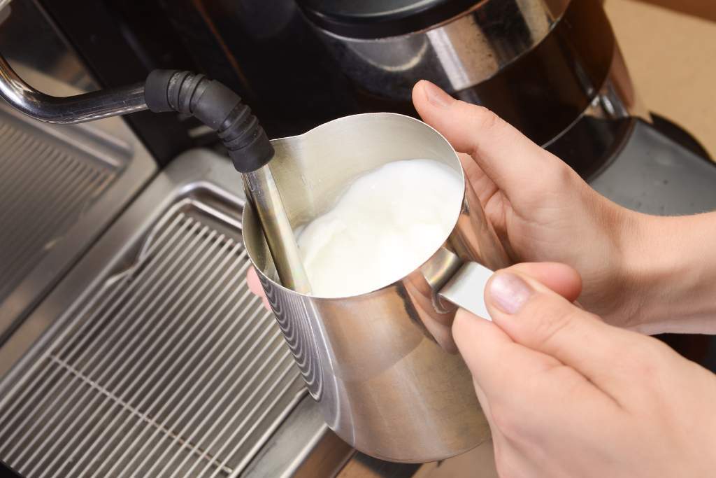 Barista steaming milk with a stainless steel jug, positioning the steam wand tip just below the surface to create smooth, glossy microfoam for coffee.