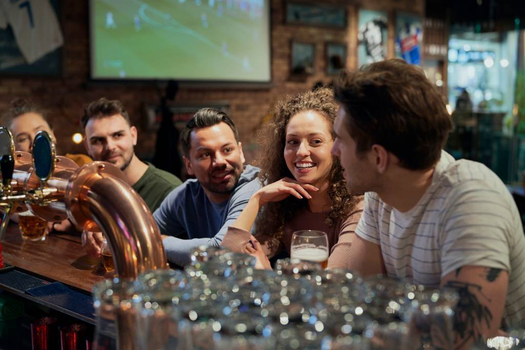 Group of young adults sitting at a bar counter, chatting and smiling while drinks are served, showing a welcoming hospitality setting.