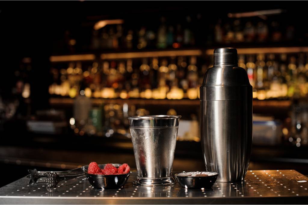 Cocktail shaker, strainer, and jigger placed neatly on a bar with fresh raspberries, illustrating bartender toolkit essentials in use.