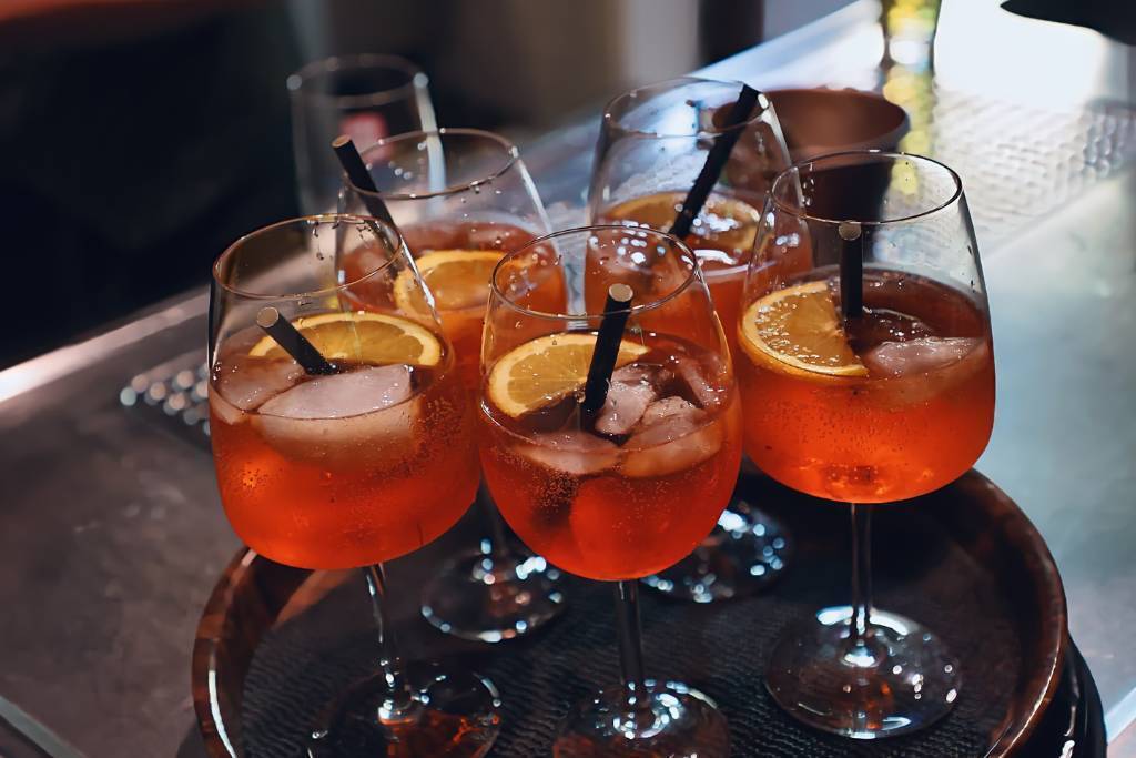 A tray of orange aperitif cocktails served in stemmed glasses, each with ice, a slice of citrus, and a black straw, arranged closely together on a bar counter, illustrating a social hospitality setting relevant to the question “How Long Does an RSA Last?”
