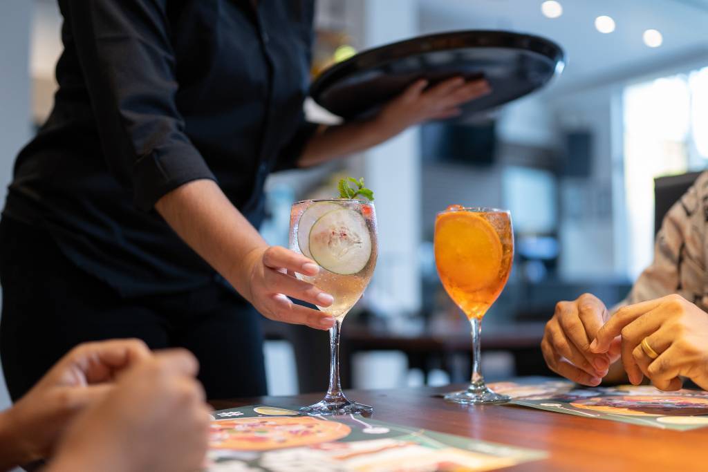 A hospitality worker serves a chilled cocktail with a citrus garnish to seated customers at a restaurant table, with other drinks visible, illustrating the benefits of getting your RSA certificate.